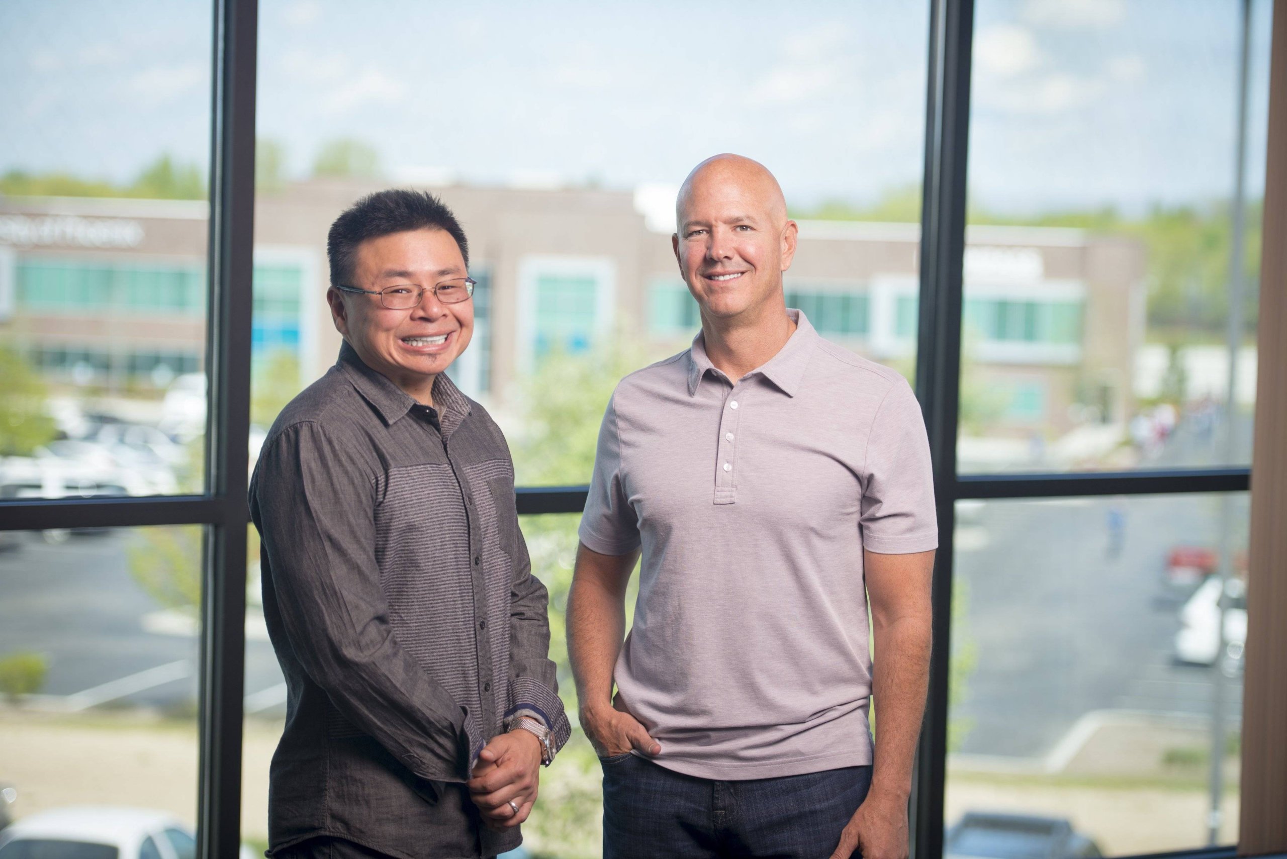 Two men stand smiling in front of a large window with a view of a parking lot and buildings