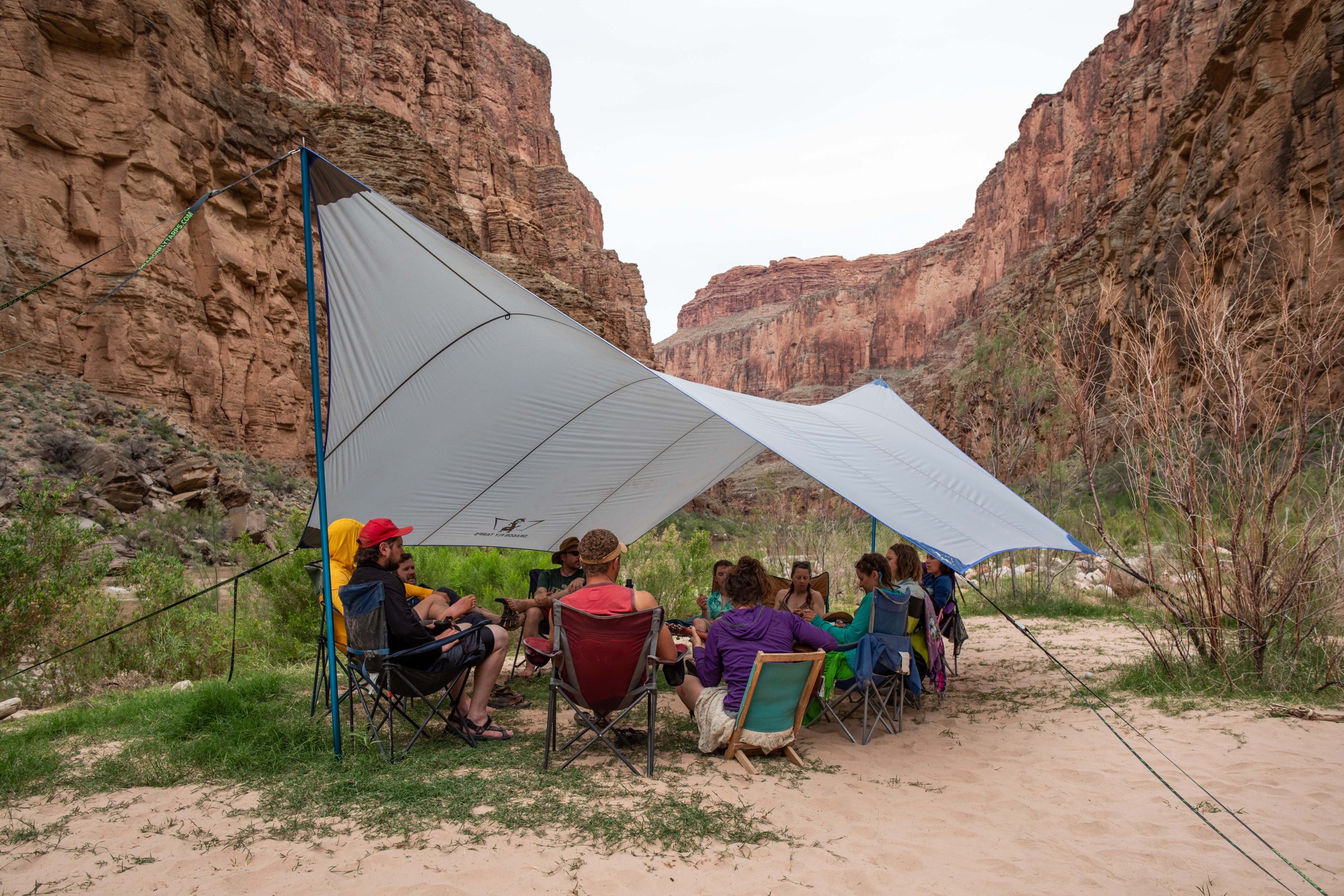 A group of people relax under a large white tarp in a desert canyon setting