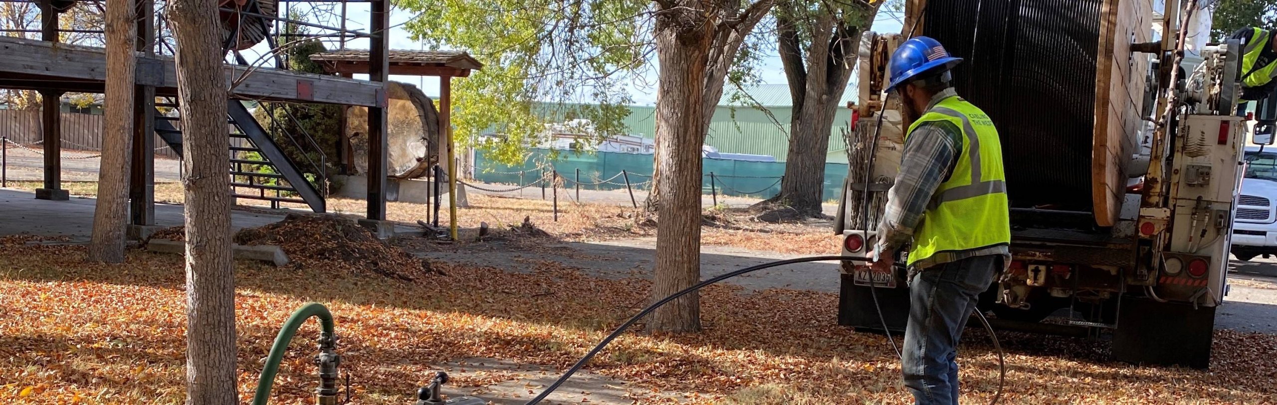 A utility worker in a high-visibility vest and hard hat operates a large cable spooling machine in a park-like setting