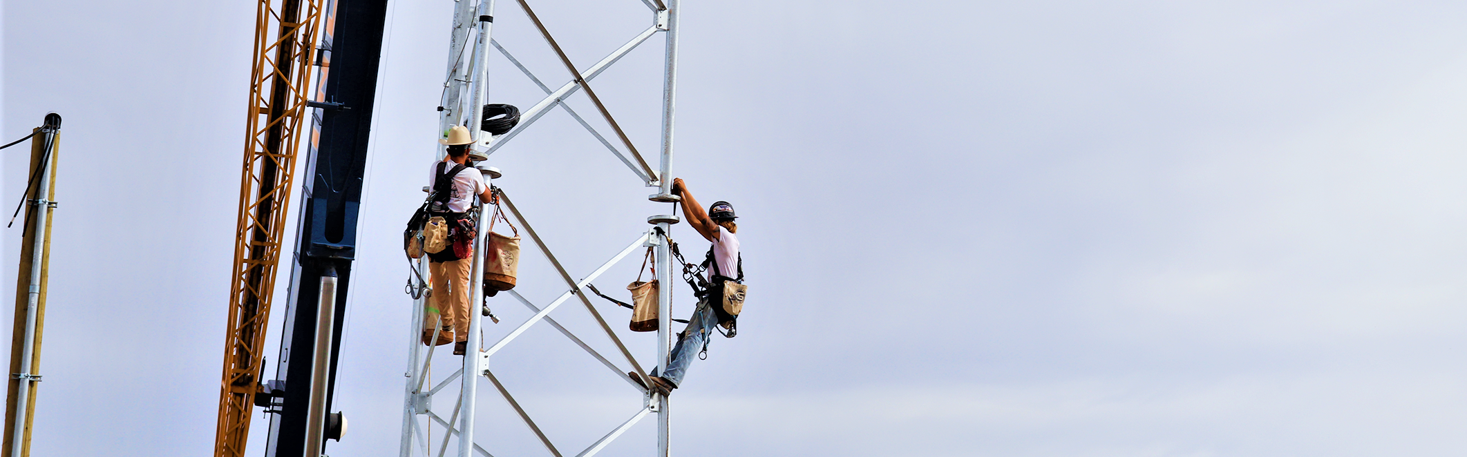 Two workers in harnesses climb a tall metal tower with a crane nearby