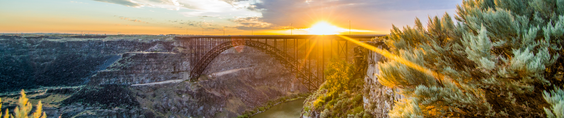 A panoramic view of a bridge spanning a deep canyon at sunset with trees and rocks