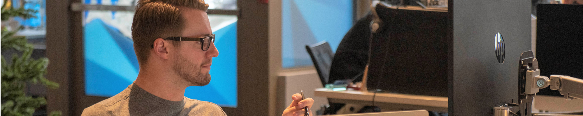 A man wearing glasses and a gray shirt is working at a desk with a computer monitor