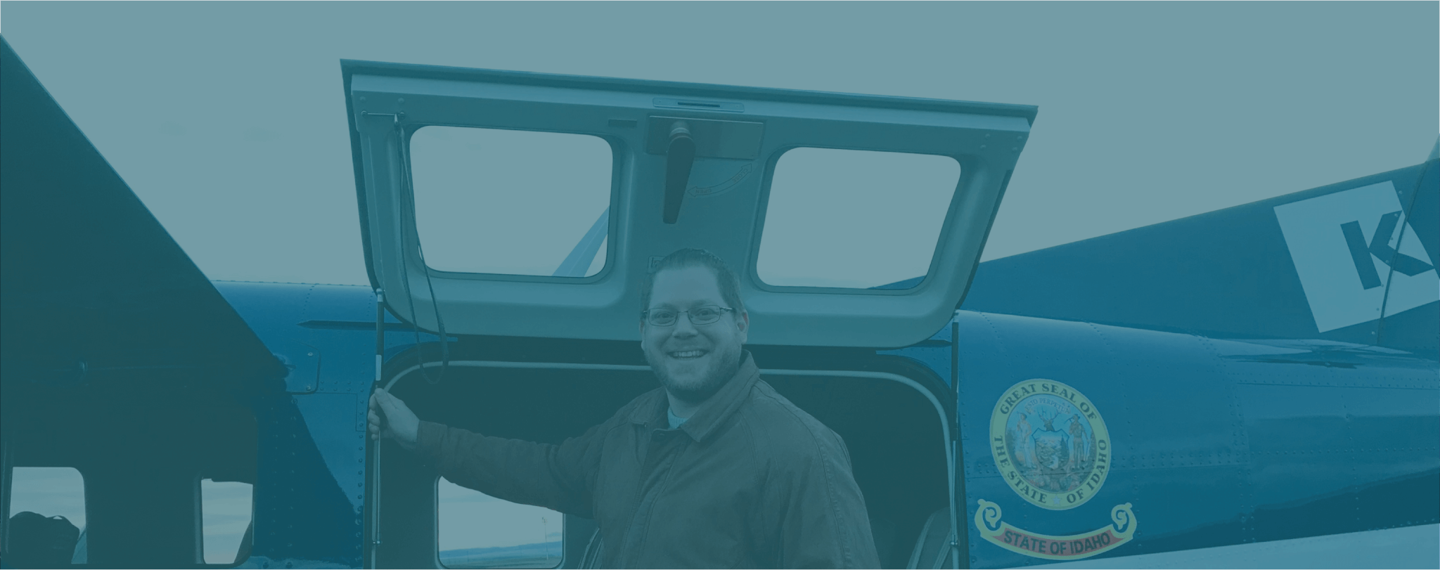 A smiling man stands in the doorway of a small airplane with the Idaho state seal