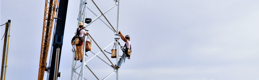 Two construction workers are climbing a tall metal tower using ropes and harnesses
