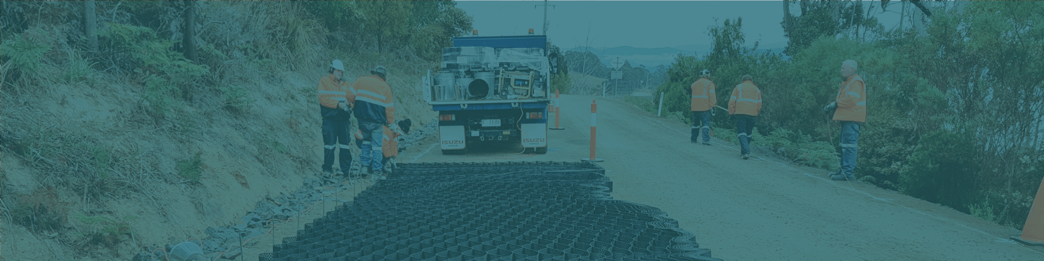 Construction workers in orange vests install a black mesh-like material on a rural road