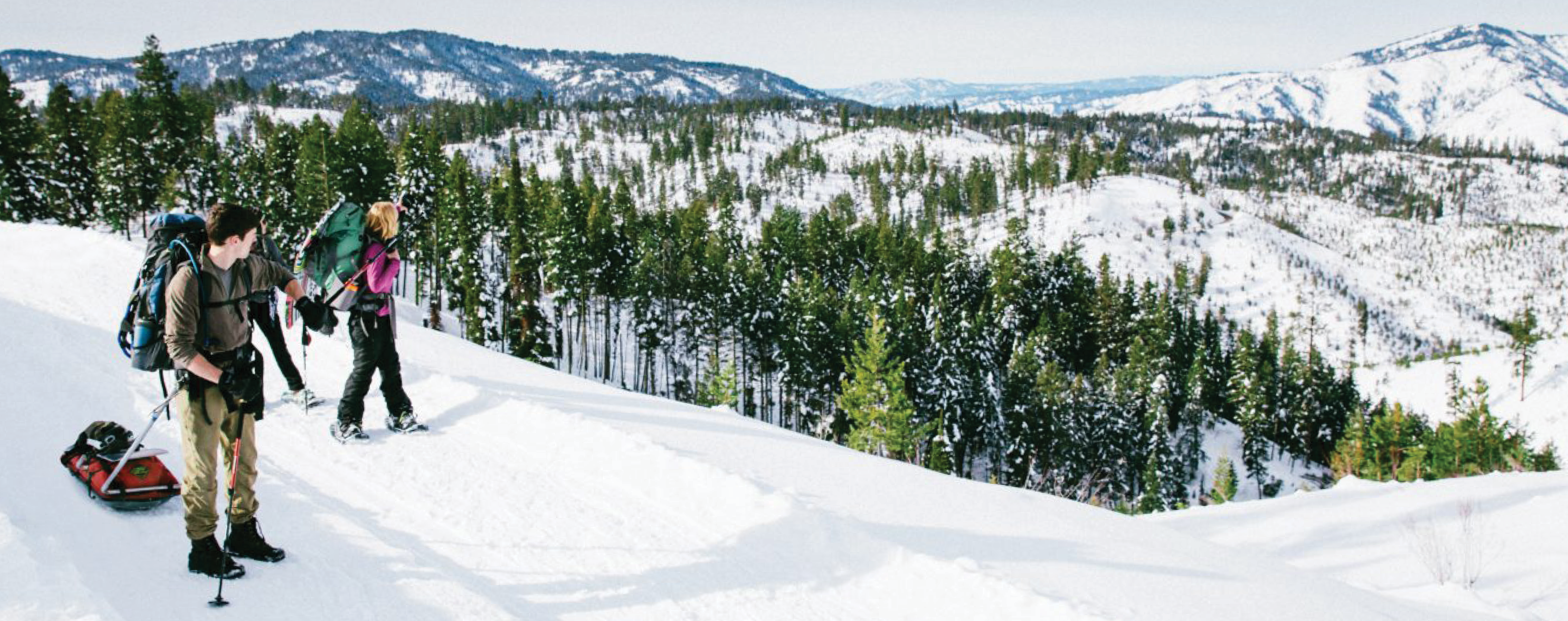 Two hikers with backpacks and snowshoes traverse a snowy mountain trail
