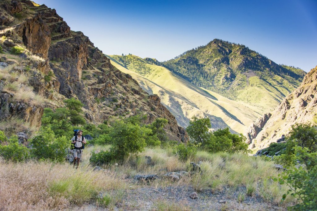 Hiker with backpack ascends rugged mountain trail through dry grass and shrubs