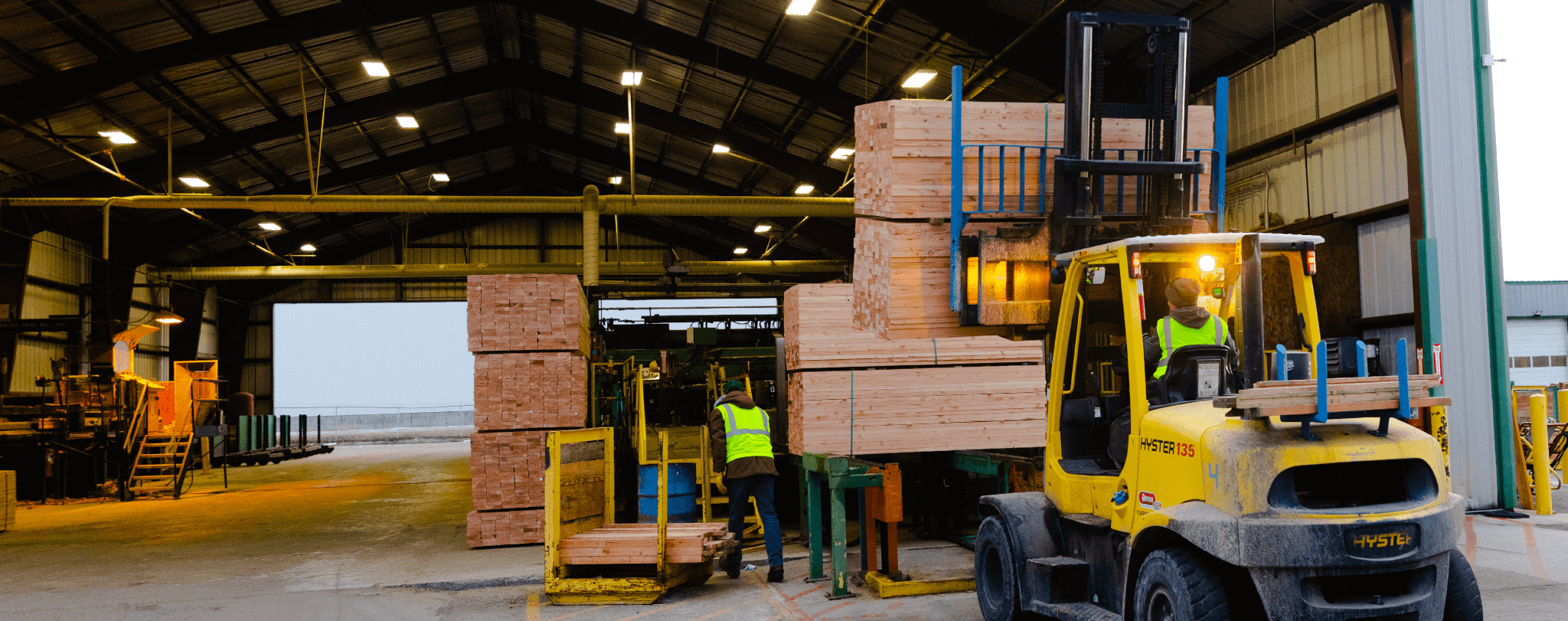 A worker in a yellow vest operates a yellow forklift to move wooden pallets in a warehouse