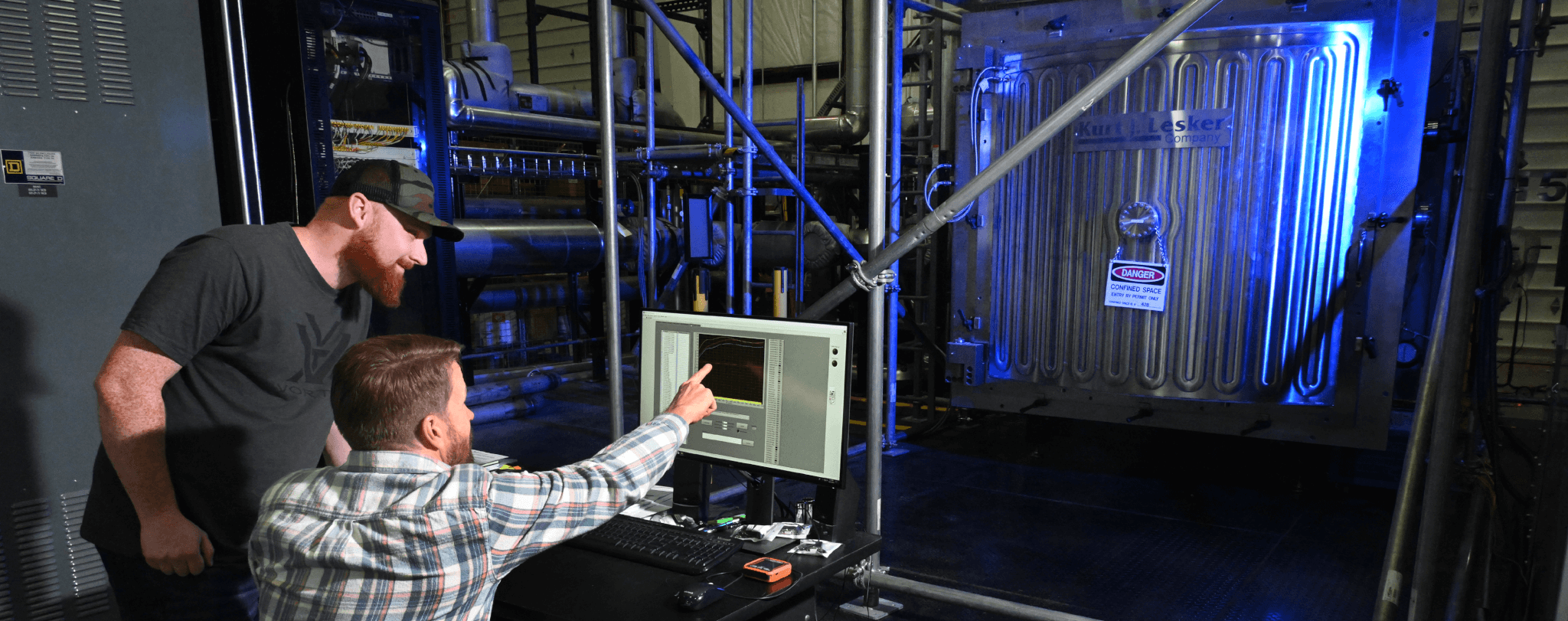 Two men in industrial setting examine computer screen with data, pointing at monitor