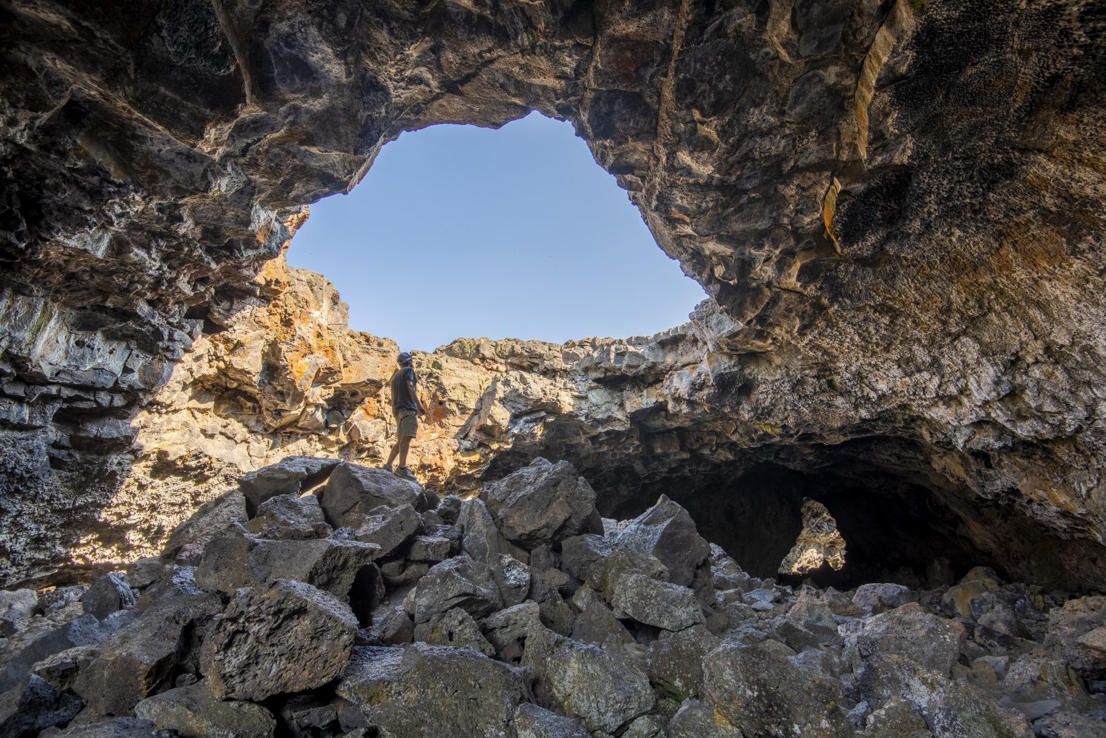 A hiker stands amidst jagged lava rocks beneath a towering volcanic cave entrance