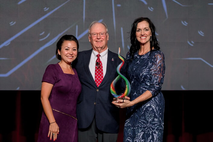 Three people, two women and a man, smiling and holding an award on a stage