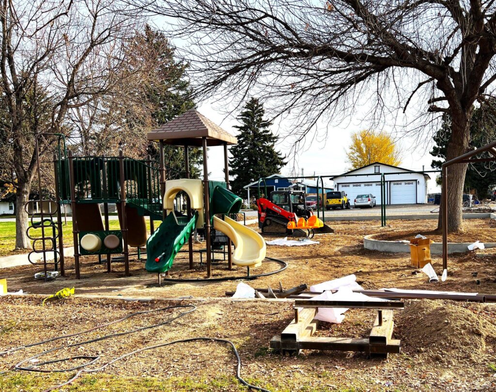 A construction site for a new playground with a partially assembled play structure, surrounded by trees and a nearby house with a garage