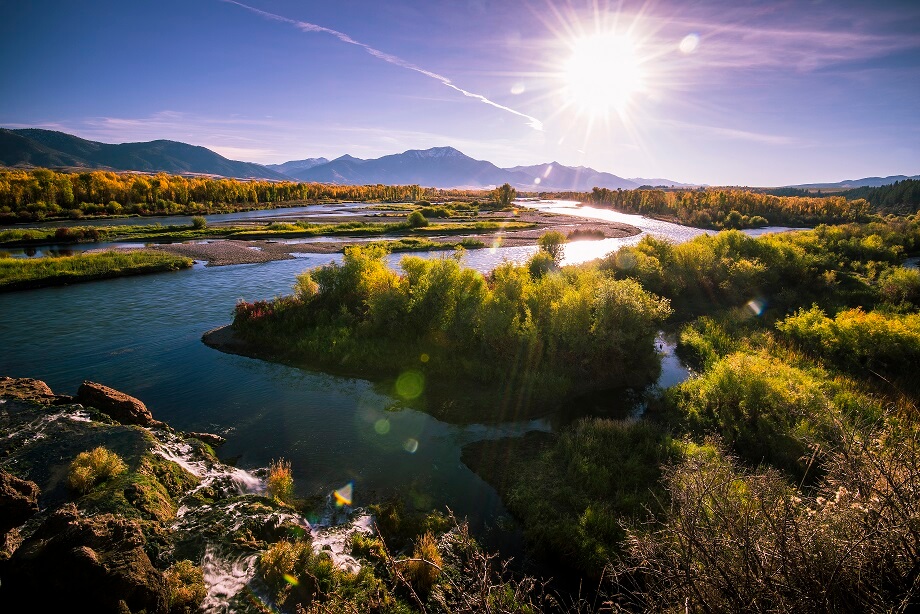 A serene landscape featuring a winding river surrounded by lush greenery and golden autumn trees, with majestic mountains in the distance under a bright, clear blue sky