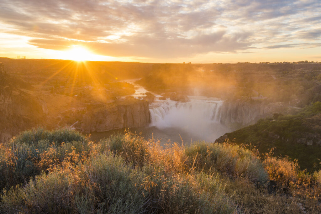 A breathtaking sunset illuminates a majestic waterfall cascading into a misty canyon, surrounded by lush greenery and golden grasses