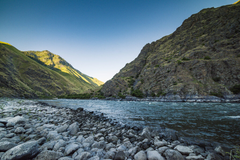 A serene river winds through a rugged canyon, flanked by towering mountains and a rocky shoreline under a clear blue sky