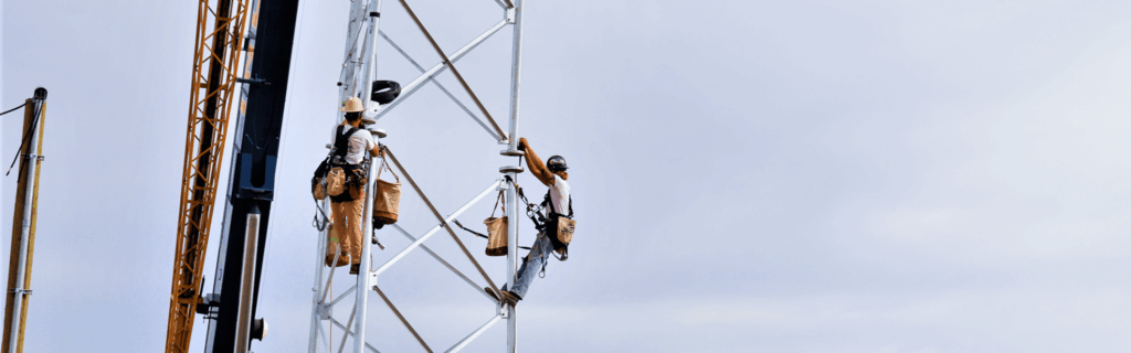 Two construction workers are scaling a tall metal tower using ropes and harnesses, with a crane visible in the background