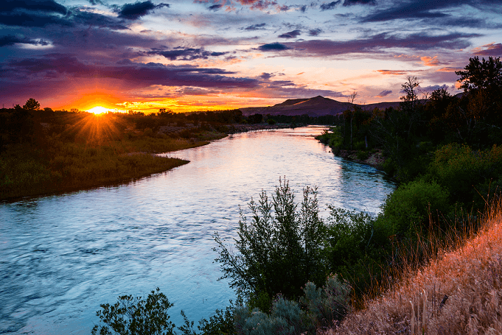 A serene river winds through a lush valley at sunset, with vibrant colors reflecting on the water's surface