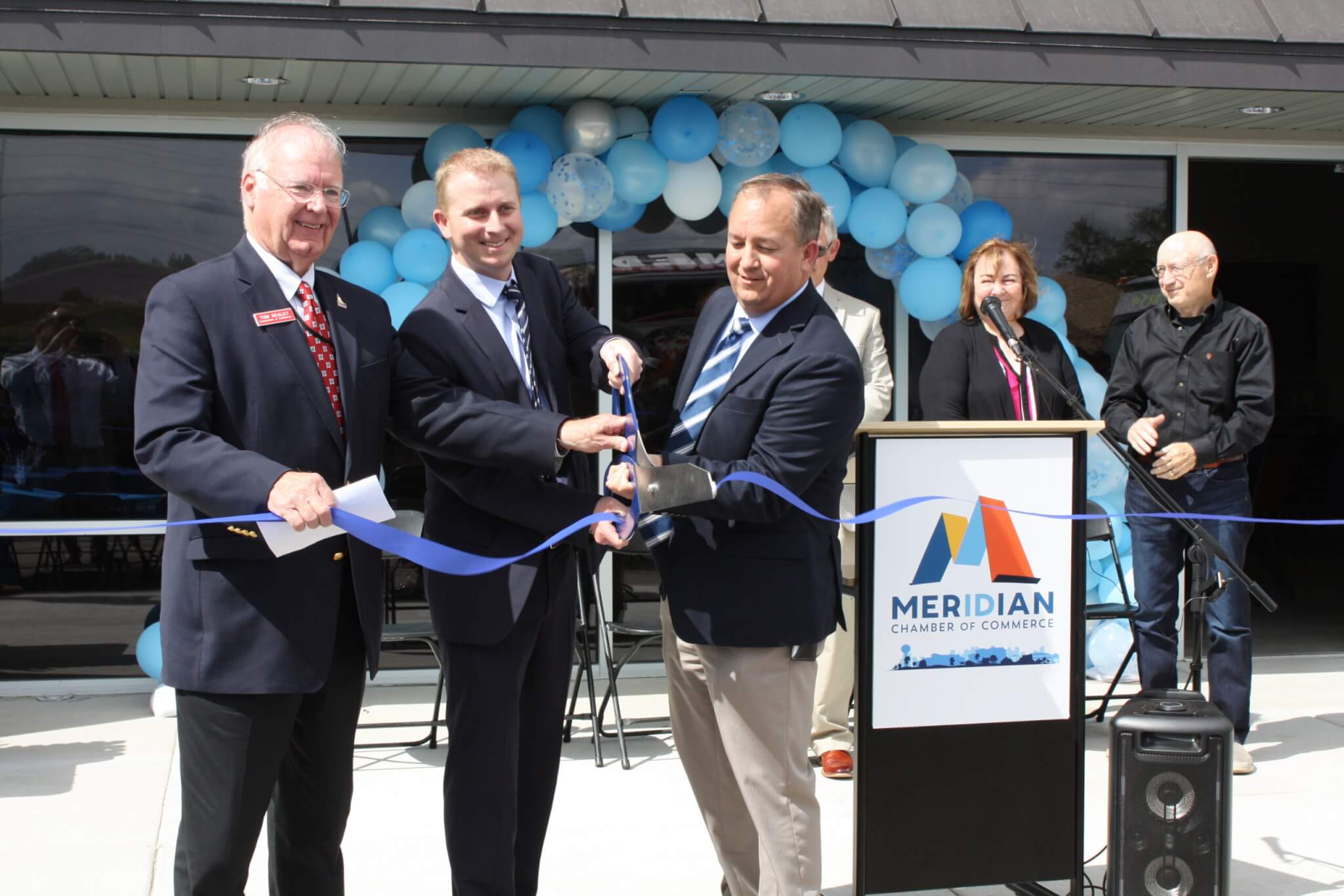 Four men in suits stand at a ribbon-cutting ceremony for the Meridian Chamber of Commerce, with balloons and a podium in the background