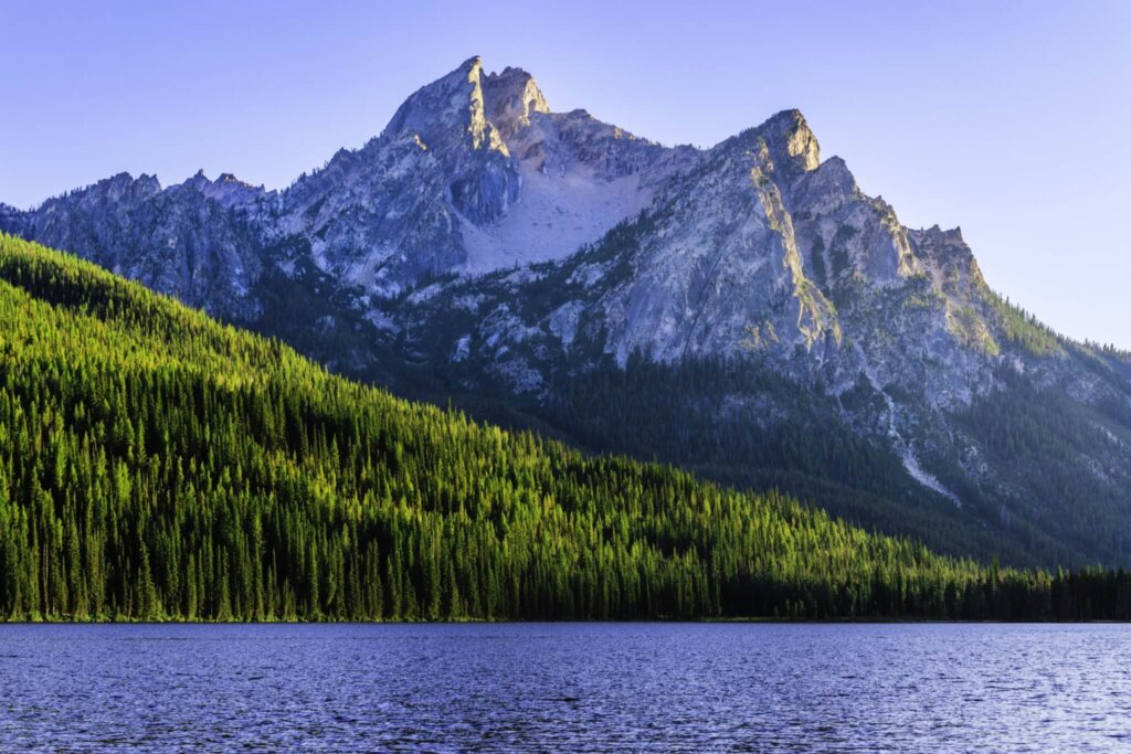Majestic mountain peaks rise above a serene lake, surrounded by lush green forests under a clear blue sky