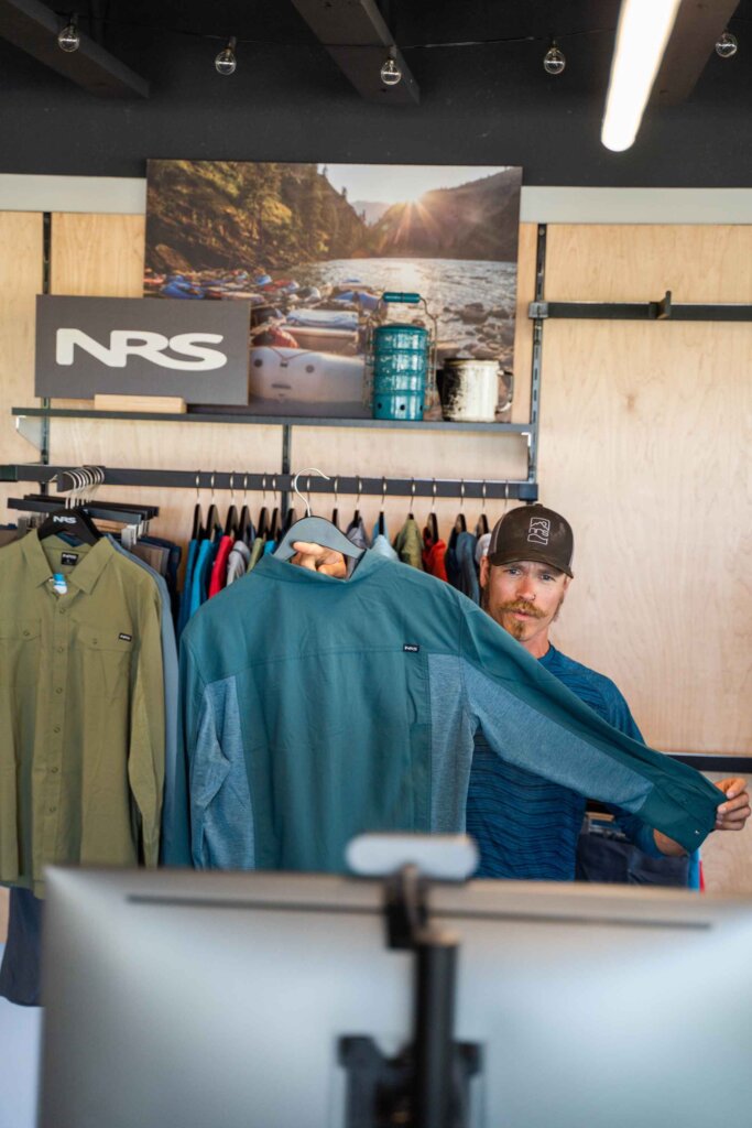 A man in a blue shirt and cap stands behind a counter, holding up a teal jacket in a clothing store with outdoor gear