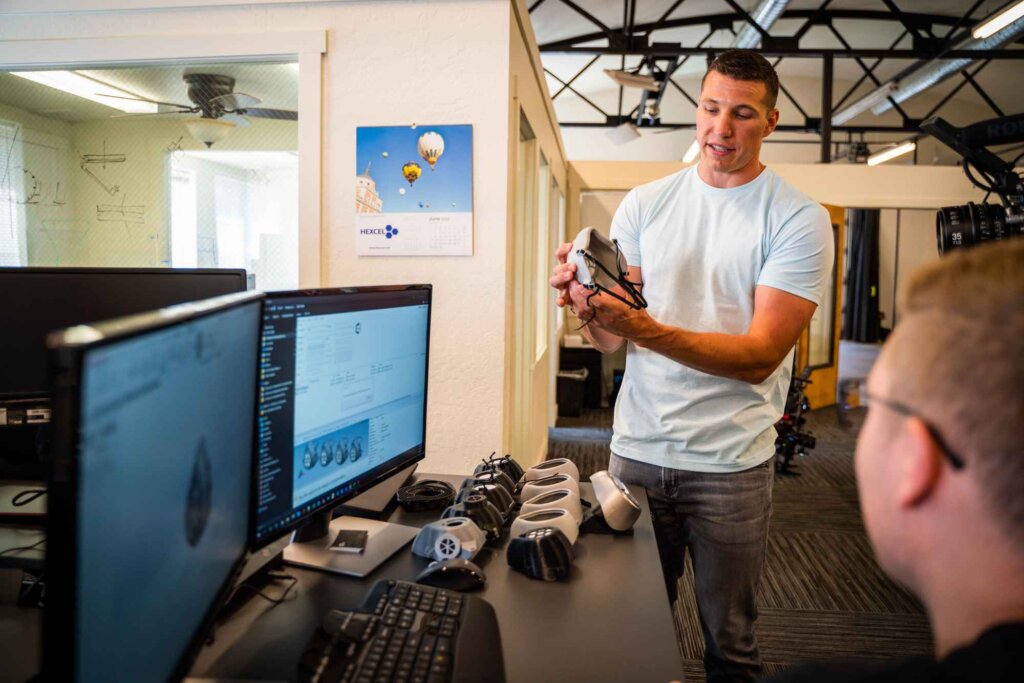 A man in a white t-shirt demonstrates a small device to a colleague in a tech office with multiple computer monitors and a calendar featuring hot air balloons