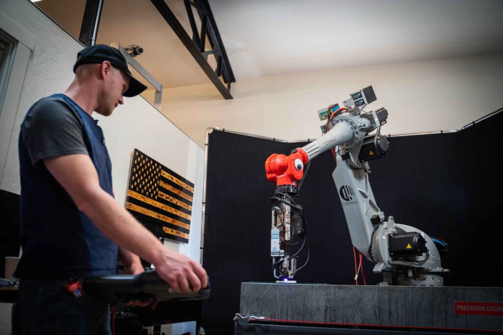 A man wearing a cap operates a control panel while observing a robotic arm with an orange gripper in a laboratory setting