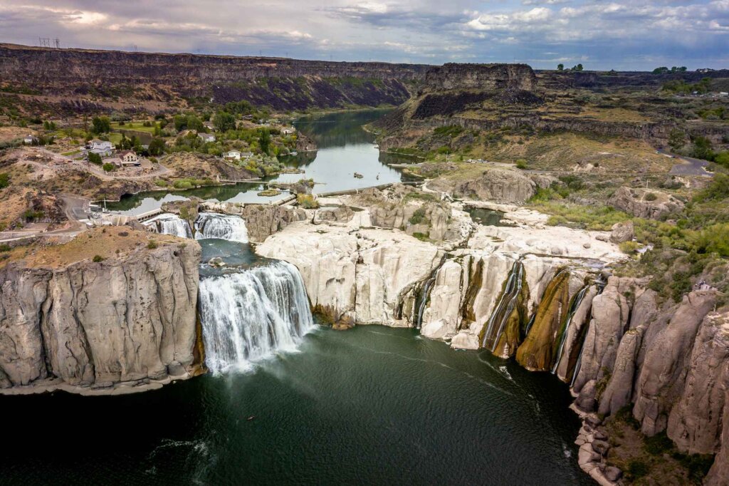 Aerial view of a majestic waterfall cascading over white cliffs into a dark river, surrounded by rugged canyon walls and a serene lake in the distance