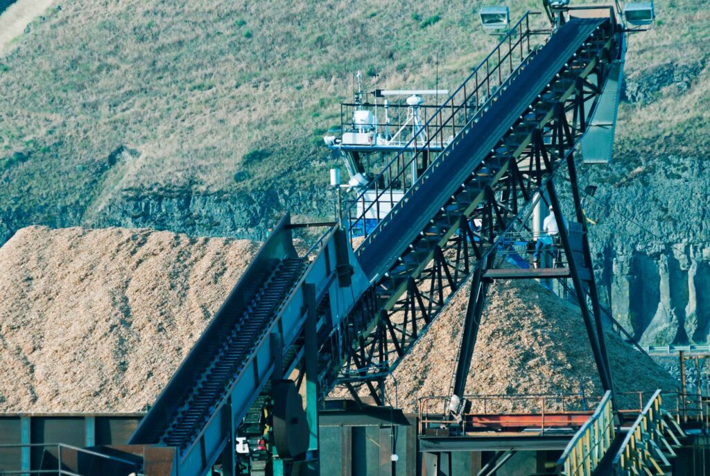A large industrial conveyor belt system transports wood chips up a steep incline towards a processing facility
