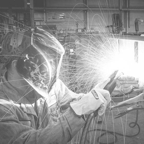 A black and white photo of a person wearing a mask, jumpsuit and work gloves metal working and making sparks fly.