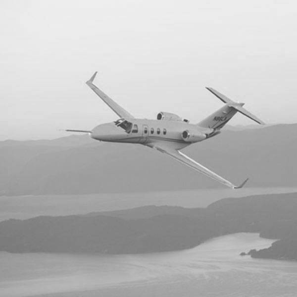 A black and white photo of a plane flying in the sky, and beneath it, a body of water and mountains.