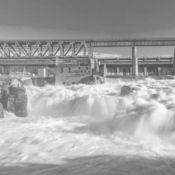 A black and white photo of water gushing through a dam, and in the background, a brick building and industrial equipment in the background.