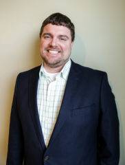A smiling man in a navy suit jacket and checkered shirt stands against a beige wall