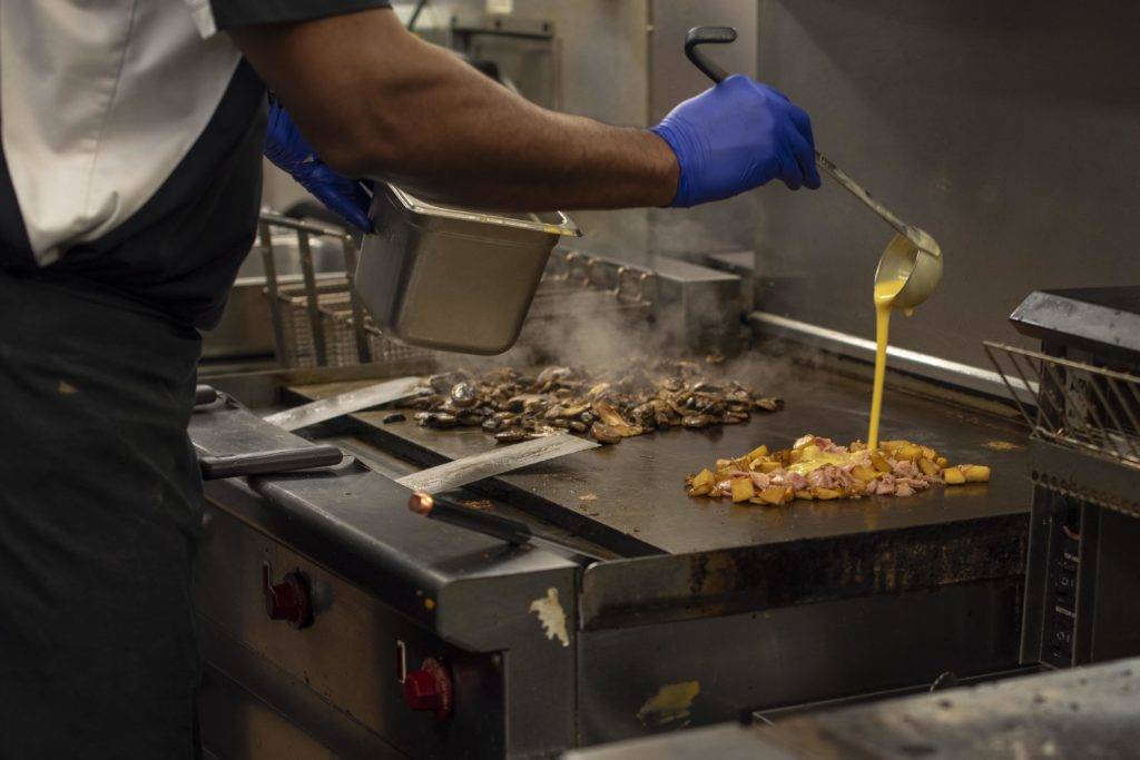 A chef in blue gloves pours yellow sauce over meat and vegetables on a hot griddle