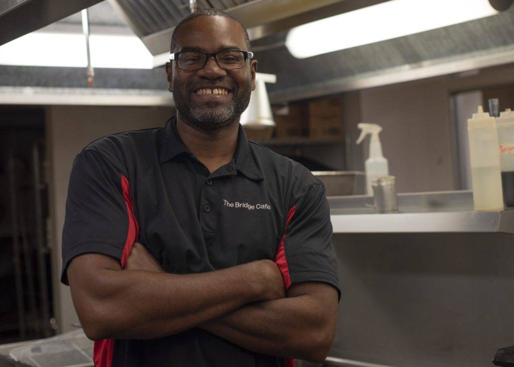 A smiling man wearing a black polo shirt with red accents stands confidently in a kitchen