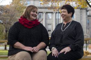 Two smiling women in black tops sit on a bench in a park with autumn trees