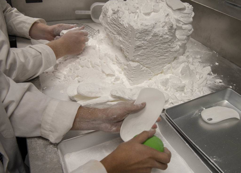 Two people in lab coats are handling white powder and objects on a metal tray