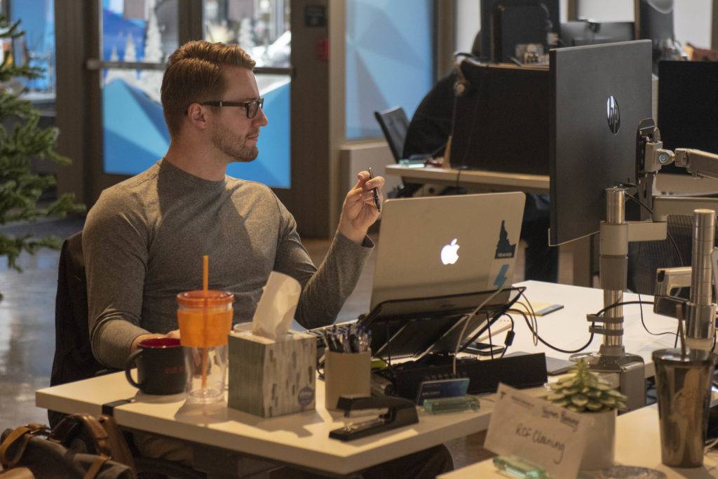 A man works at a desk with a laptop, monitor, and various office supplies