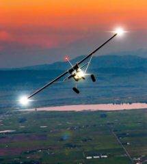 Small airplane flying low over a vast landscape at sunset with landing lights on