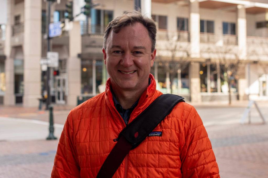 A smiling man wearing an orange jacket stands outdoors in an urban setting