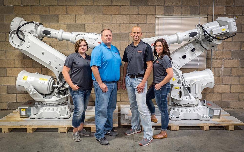 Four people stand in front of two large industrial robotic arms in a warehouse