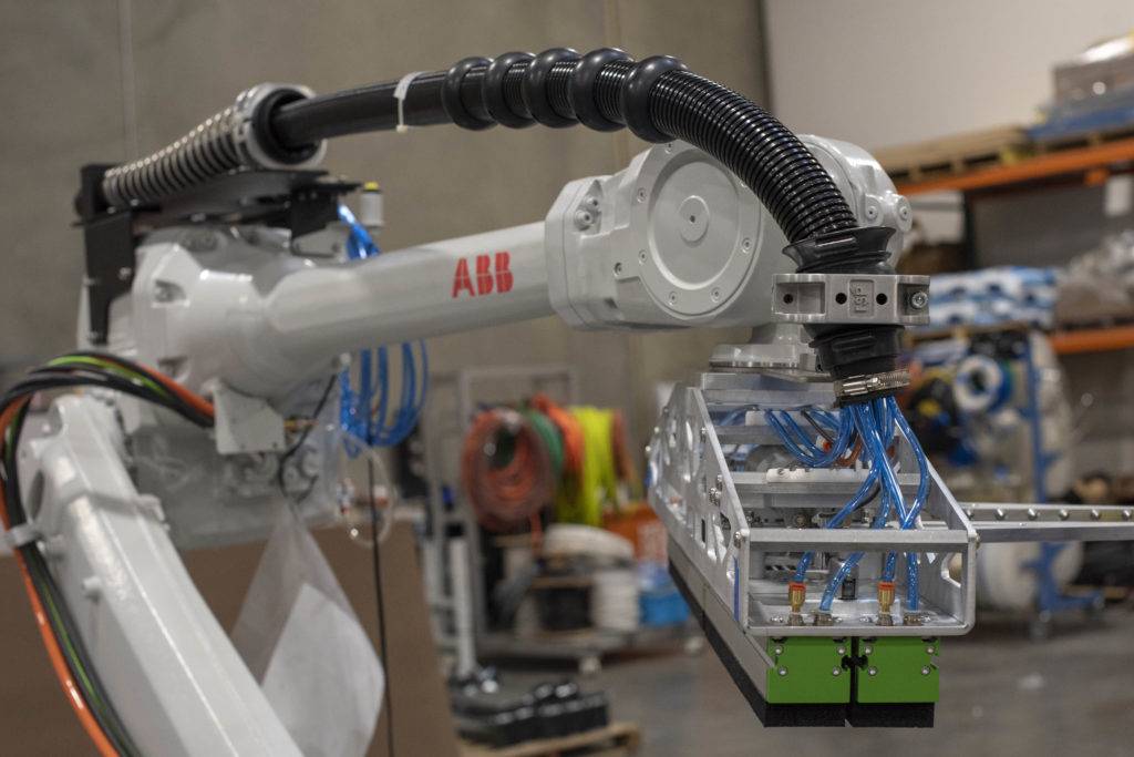 A white ABB robotic arm with blue and black tubing extends over a conveyor belt in a warehouse