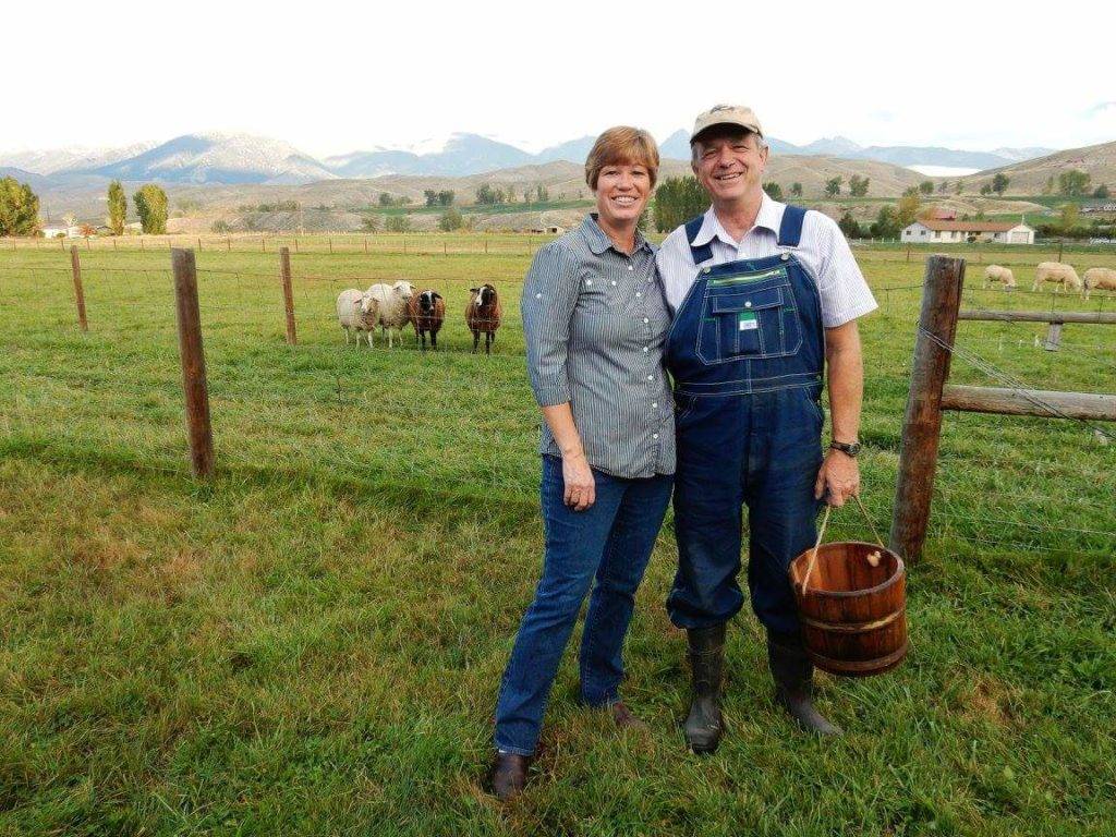 A smiling farmer and his wife stand in a grassy field with sheep and mountains in the background
