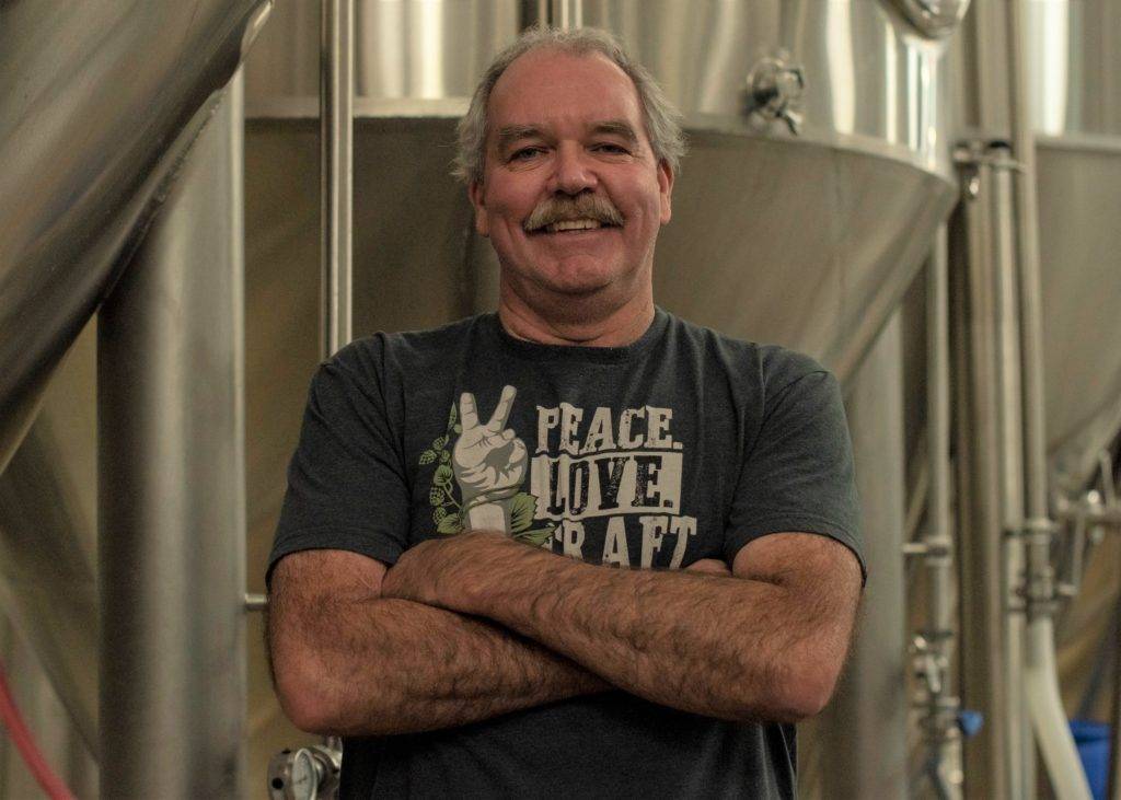 A man with a mustache stands confidently in a brewery, arms crossed, wearing a peace-themed t-shirt