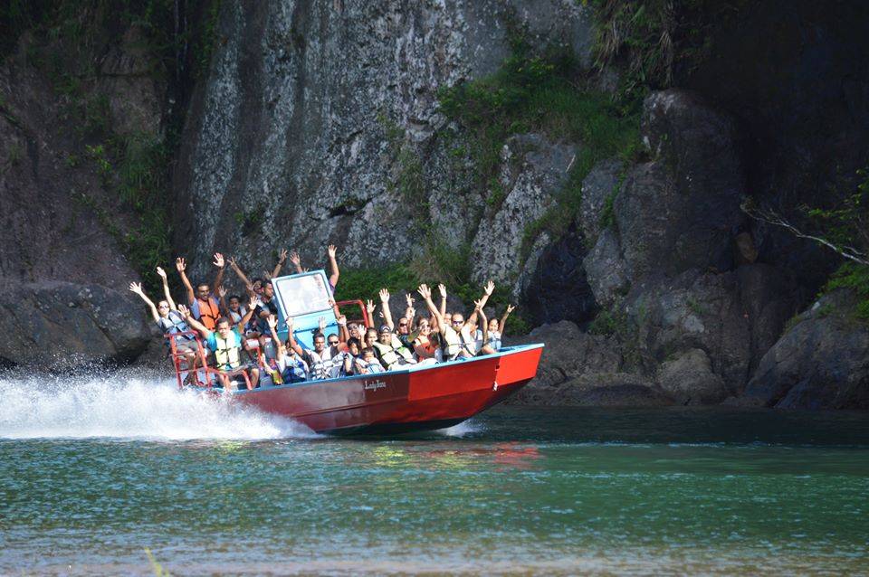 A group of people on a red boat wave excitedly as they speed past rocky cliffs
