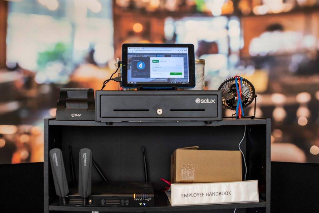 A black shelf holds a tablet, cash register, fan, and employee handbook in a blurred indoor setting