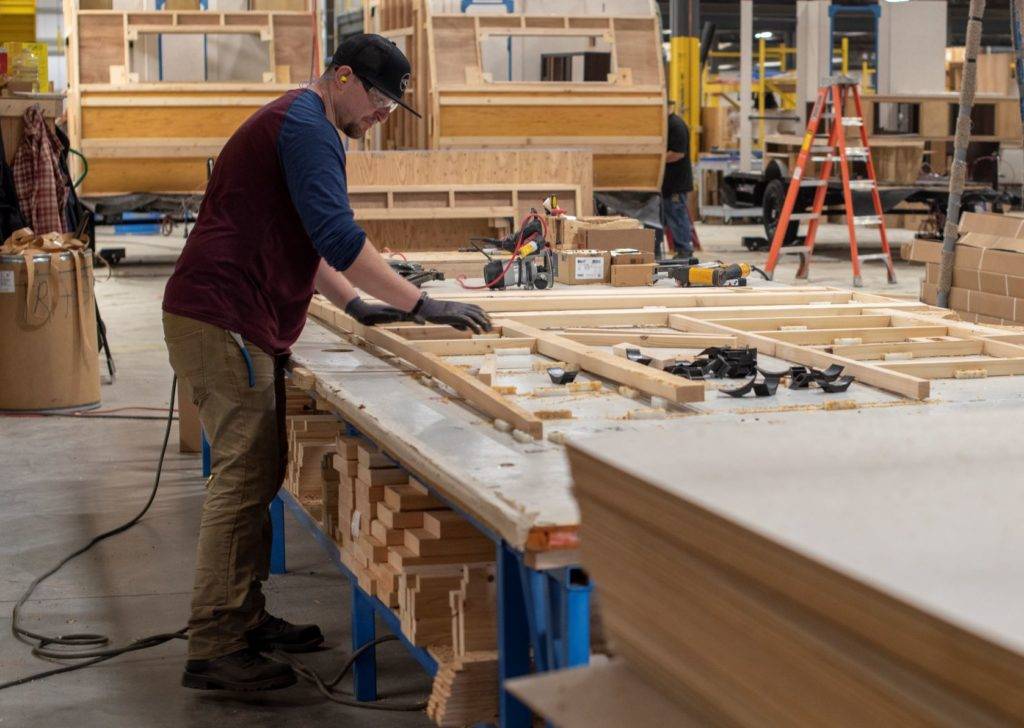 A man wearing safety gear assembles wooden frames on a long workbench in a factory