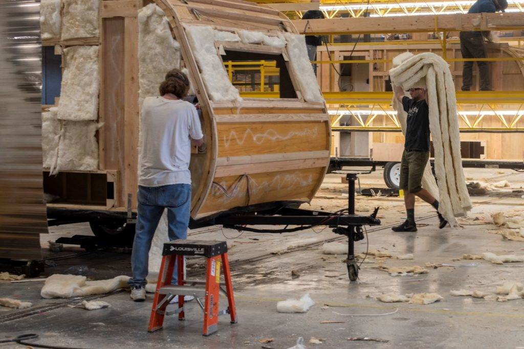 Two workers assemble a wooden structure with insulation in a large workshop