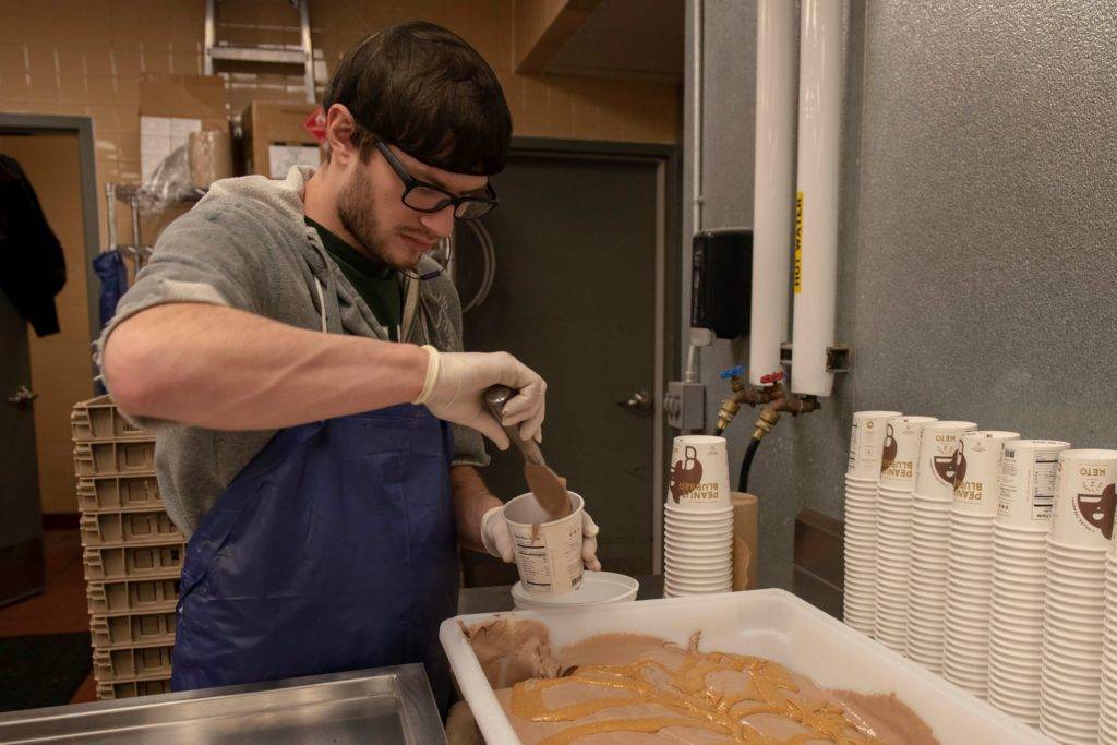 A worker in a kitchen pours a thick, brown substance from a cup into a tray while wearing gloves and an apron
