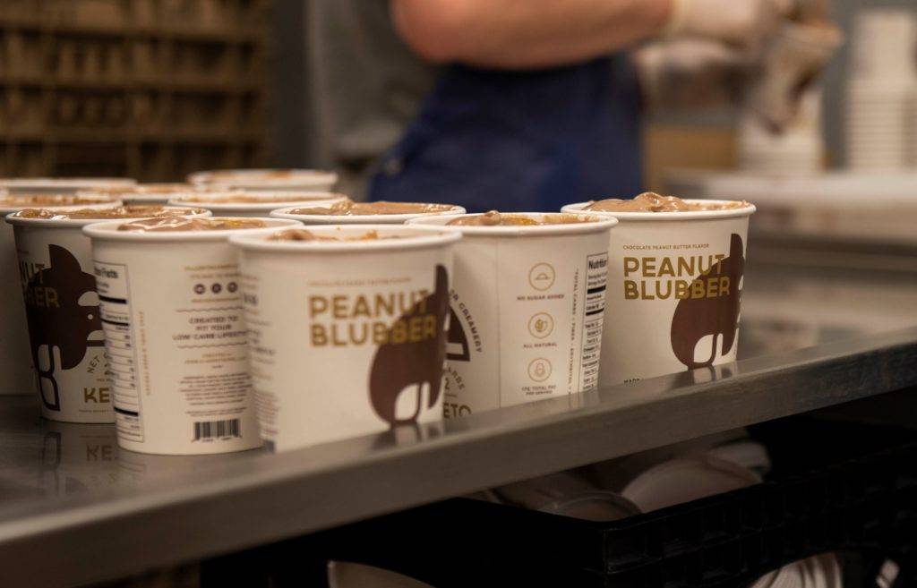 A row of white cups with brown labels labeled 'Peanut Blubber' filled with brown substance