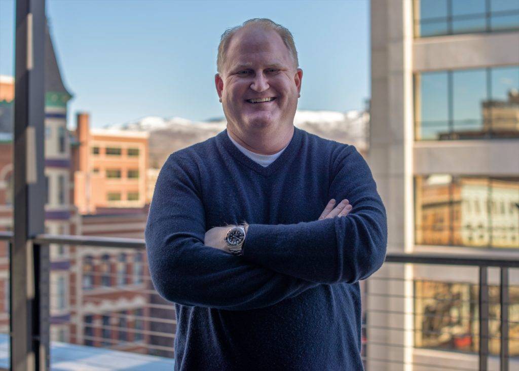 A man in a dark sweater stands confidently on a rooftop with city buildings and mountains in the background